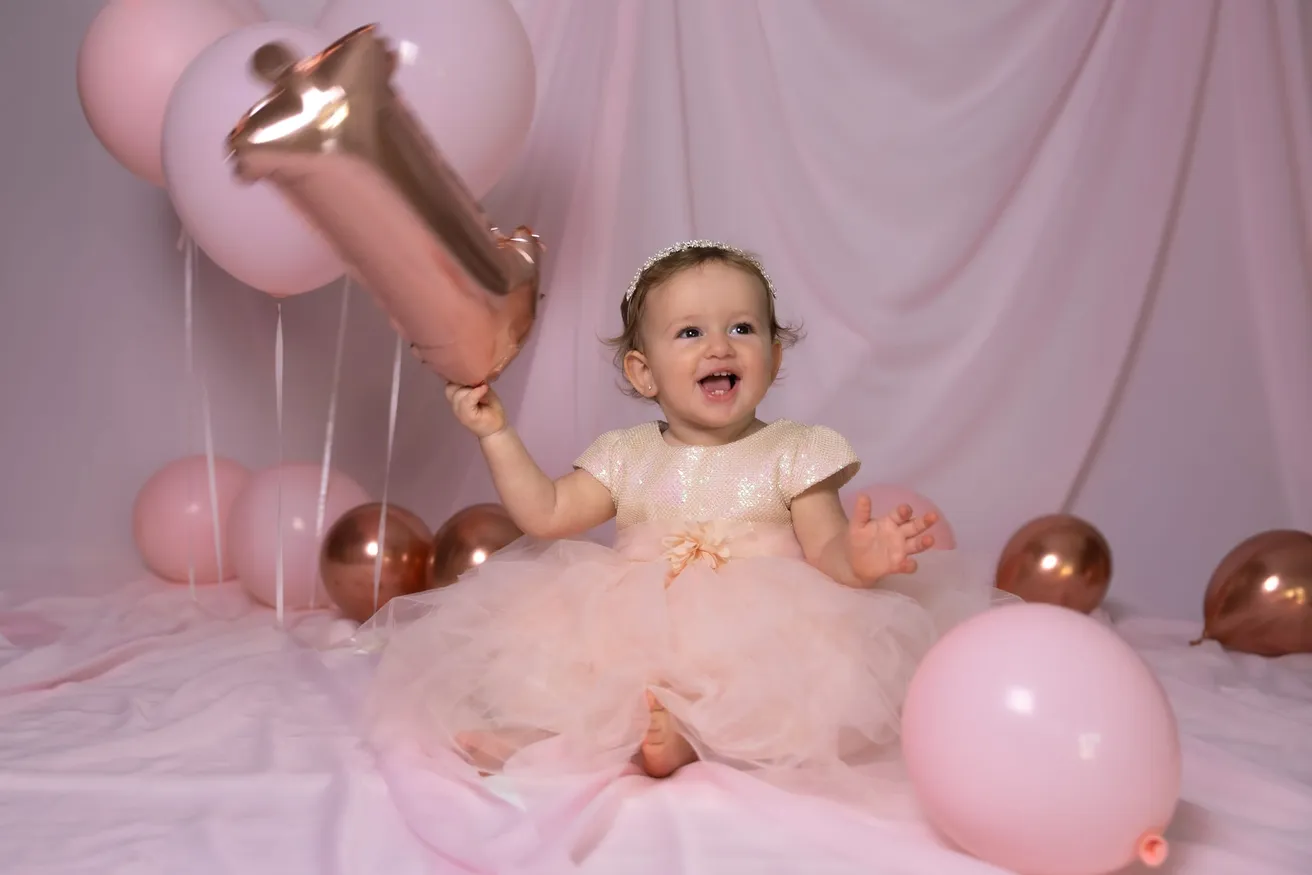 Une petite fille en robe de tulle rose rit, un ballon « 1 » à la main, au milieu des ballons roses et cuivrés