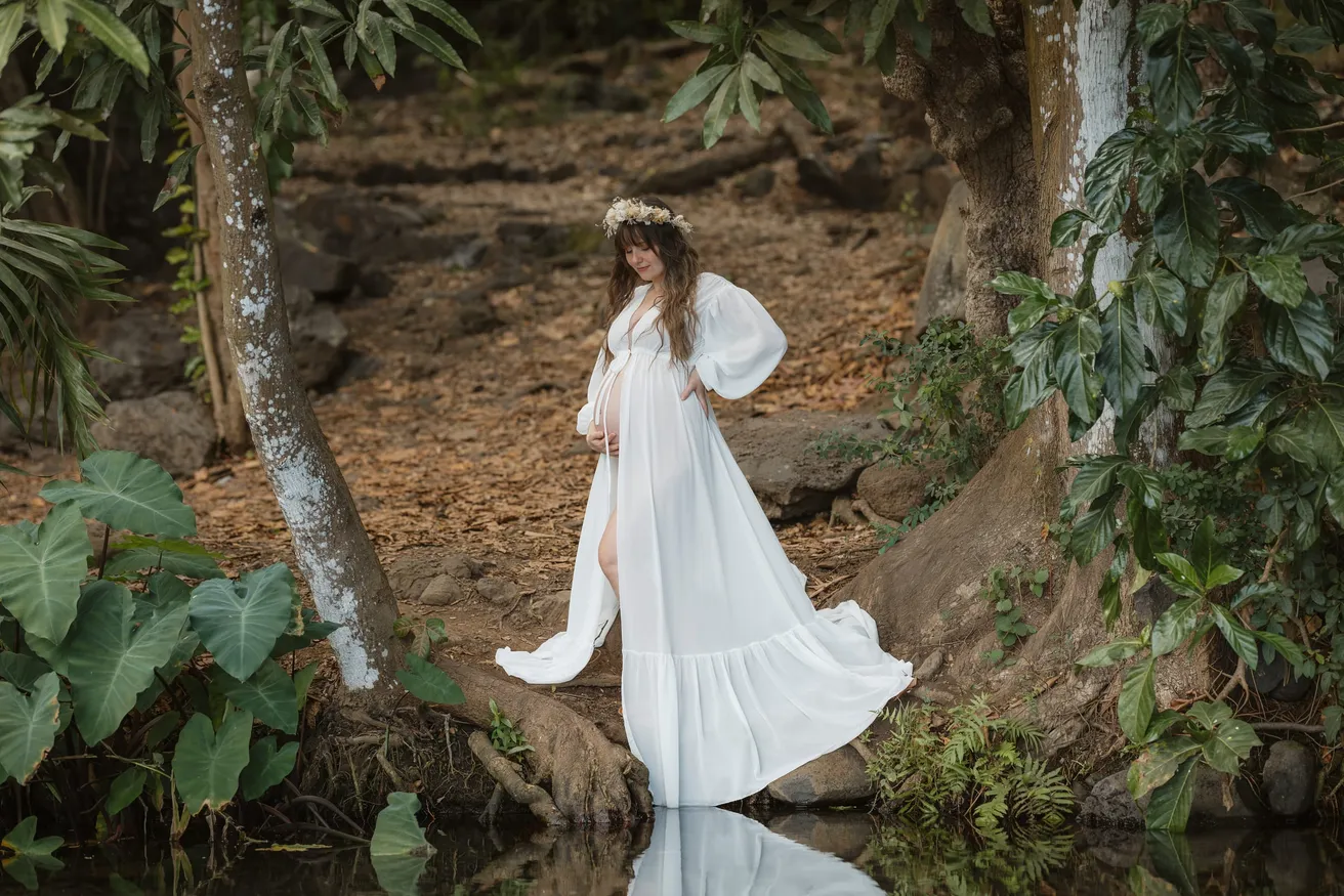 Future maman en longue robe blanche au bord d’un bassin, sous les arbres d’un sous-bois