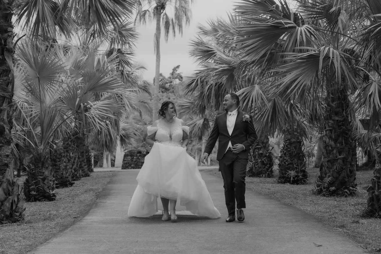 En noir et blanc, les mariés marchent dans une allée de palmiers, la mariée relève sa robe