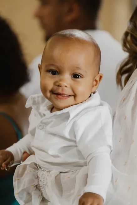 Un bébé en tenue blanche sourit à l’objectif, porté dans les bras