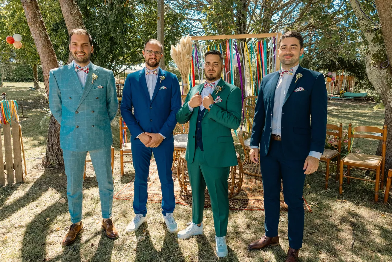 Le marié en costume vert pose avec trois témoins en costumes bleus devant une arche de rubans
