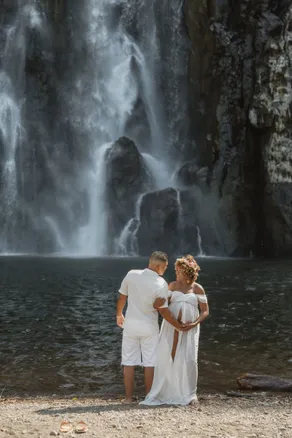 Un couple de dos face à une haute cascade, main dans la main au bord du bassin