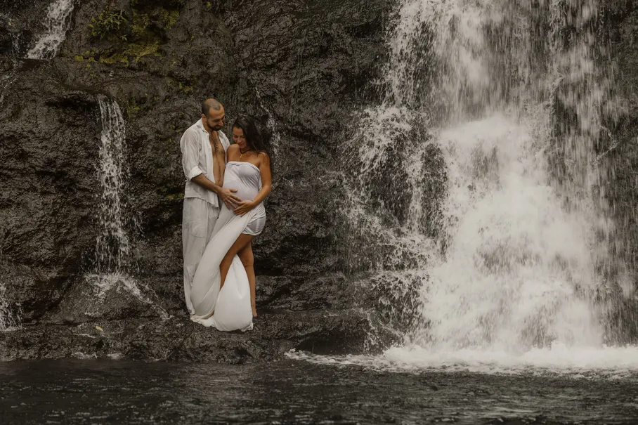 Un couple enlacé devant une cascade, la future maman drapée d’un voile blanc