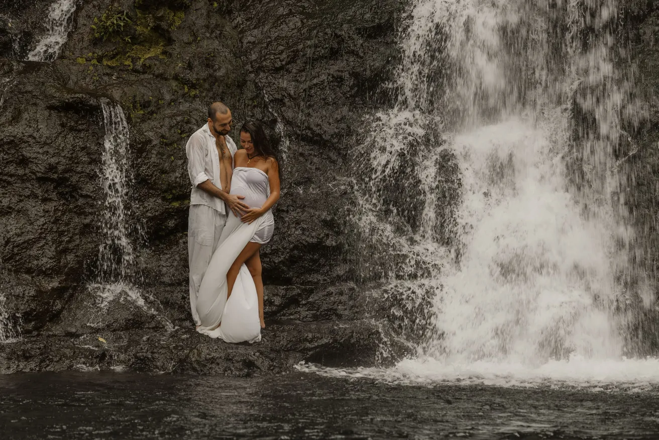 Un couple enlacé devant une cascade, la future maman drapée d’un voile blanc