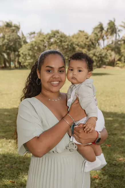 Une maman souriante tient son bébé en blanc dans ses bras, sur une pelouse bordée d’arbres