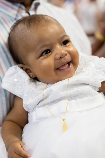 Un bébé en tenue blanche rit aux éclats, une chaîne dorée autour du cou