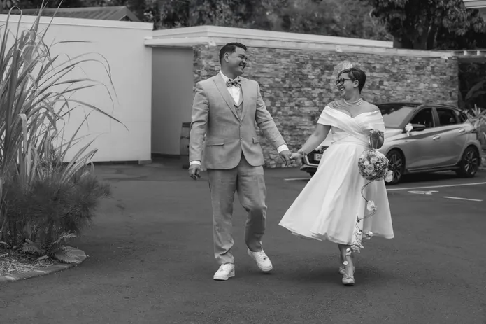En noir et blanc, les mariés marchent main dans la main, la mariée en robe courte