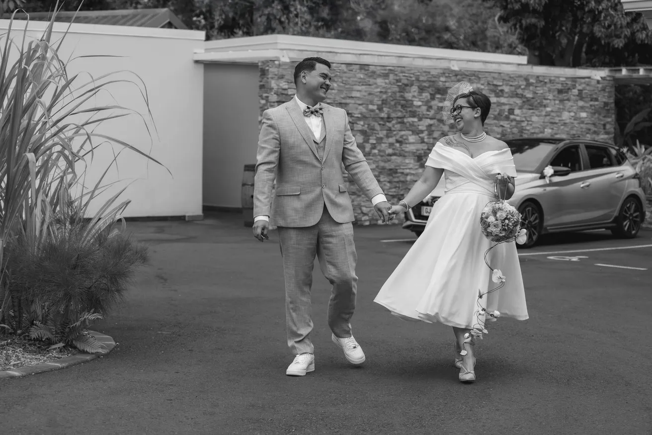 En noir et blanc, les mariés marchent main dans la main, la mariée en robe courte