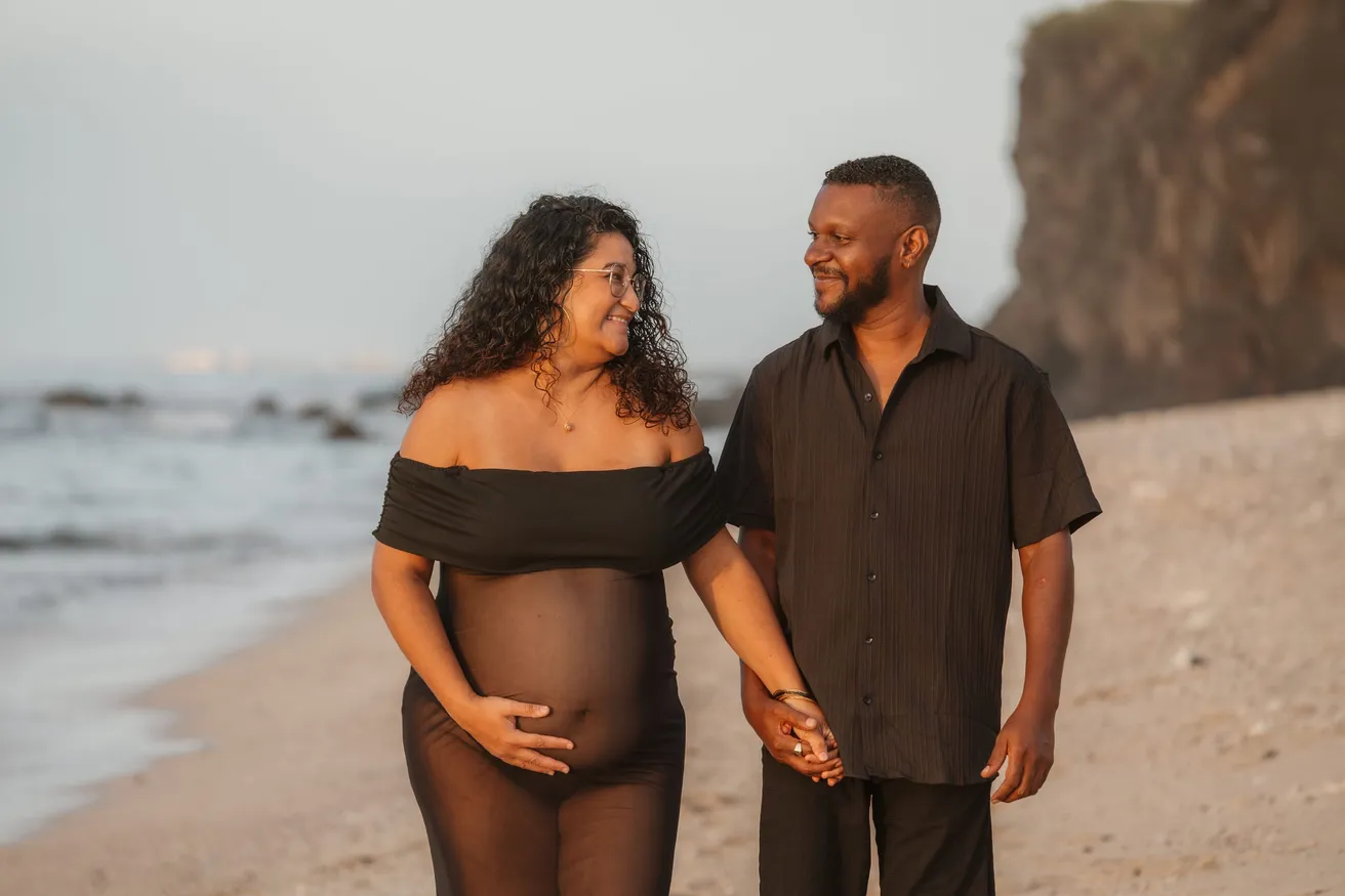 Un couple main dans la main sur la plage, la future maman en robe noire transparente