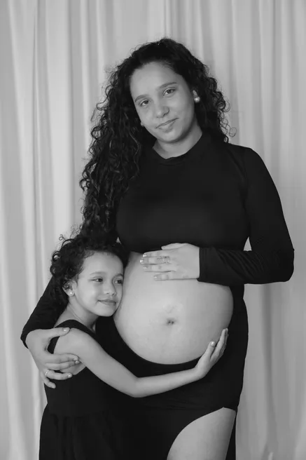 En noir et blanc, future maman en noir et sa fille qui l’enlace, devant un rideau clair