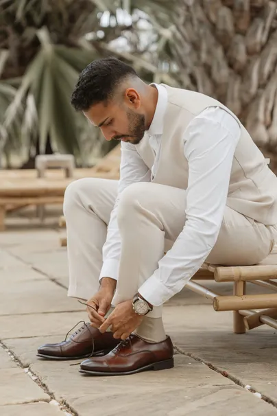 Le marié en gilet beige lace ses chaussures de cuir, assis sur un banc de bambou