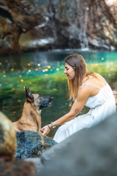 Au bord d’un bassin d’eau verte, une femme en robe blanche se penche vers son berger malinois assis sur un rocher