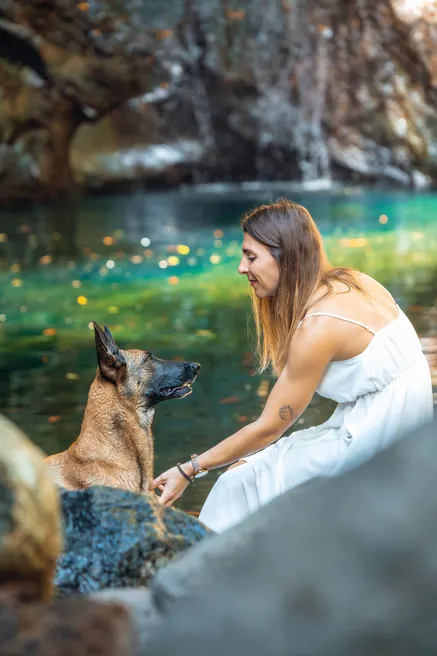 Au bord d’un bassin d’eau verte, une femme en robe blanche se penche vers son berger malinois assis sur un rocher