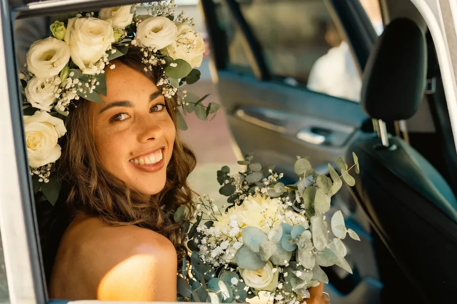La mariée en couronne de fleurs sourit depuis la portière d’une voiture