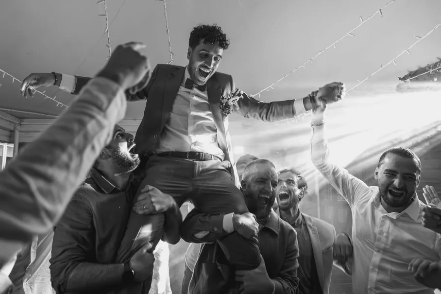 En noir et blanc, le marié porté à bras levés par ses amis, en pleine fête