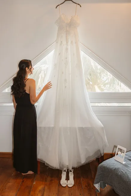 Une femme en robe noire regarde par la fenêtre, près de la robe de mariée suspendue