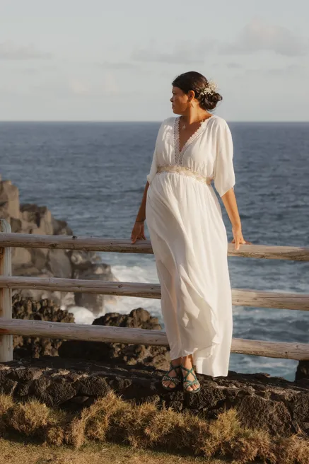 La mariée en robe blanche fluide appuyée à une barrière de bois au-dessus de l’océan