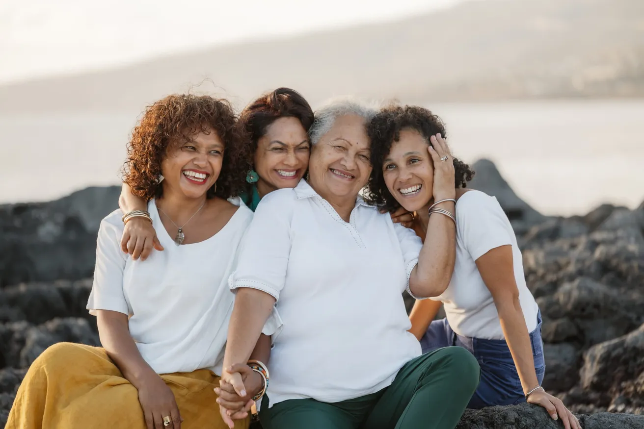 Trois générations de femmes, bras dessus bras dessous, sourient sur les rochers noirs du bord de mer