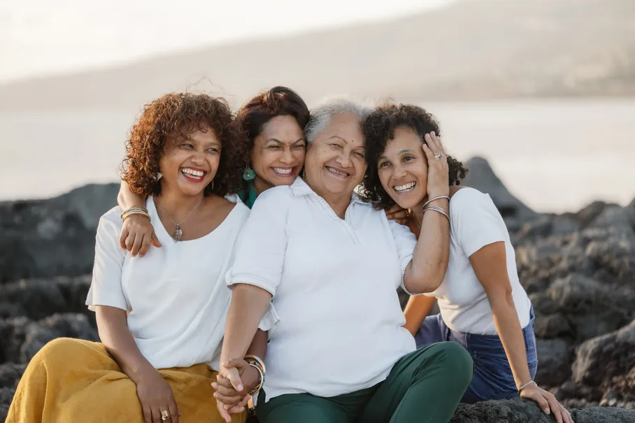 Trois générations de femmes, bras dessus bras dessous, sourient sur les rochers noirs du bord de mer