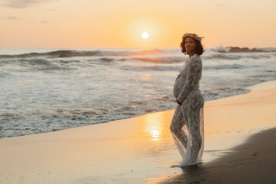 Future maman en robe de dentelle sur la plage, face au soleil couchant sur la mer
