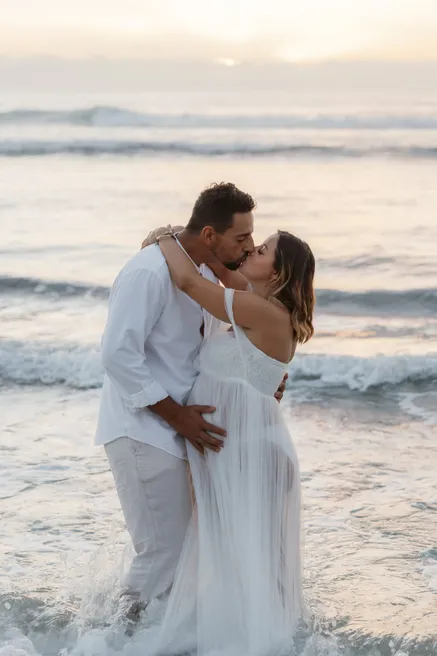 Un couple s’embrasse dans les vagues, la future maman en robe blanche transparente