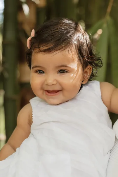 Une petite fille aux cheveux bruns sourit en robe blanche, devant des tiges de bambou