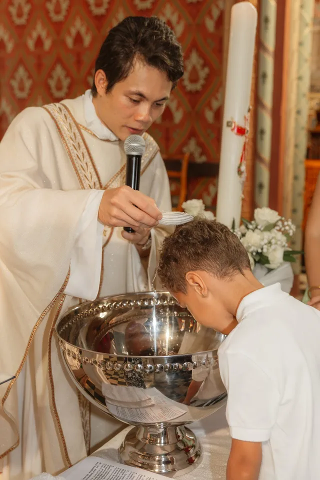 Un prêtre verse l’eau du baptême sur la tête d’un petit garçon penché au-dessus d’une vasque argentée