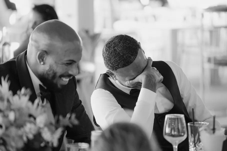 En noir et blanc, deux hommes rient à table pendant la réception, l’un se cache le visage