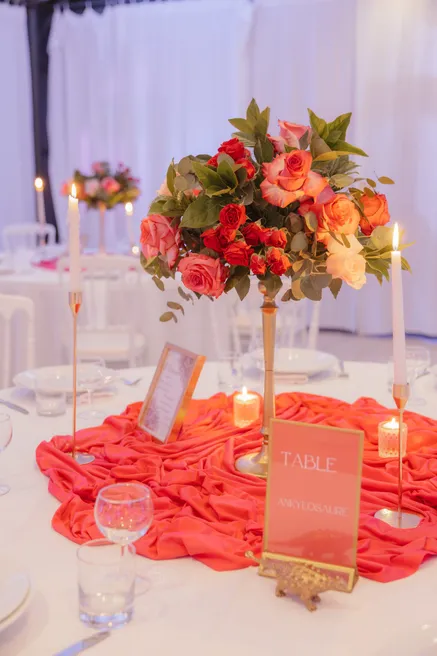 Centre de table de roses rouges sur un drapé corail, bougies et chevalet de numéro de table