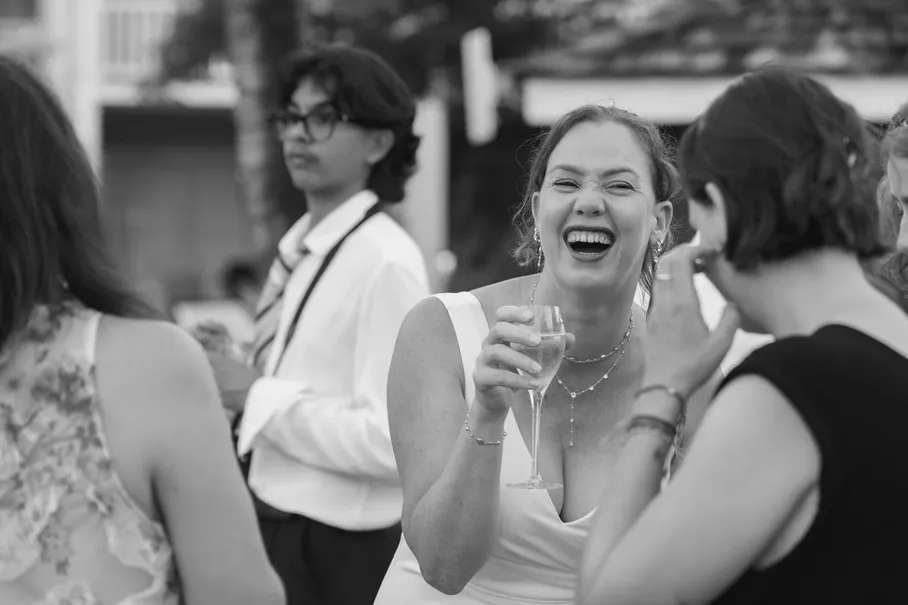 En noir et blanc, une invitée rit aux éclats, coupe de champagne à la main