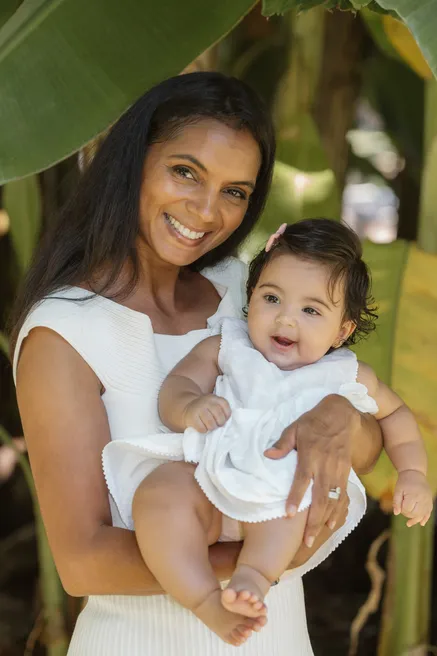 Une mère sourit en portant sa petite fille en robe blanche, devant des feuilles de bananier