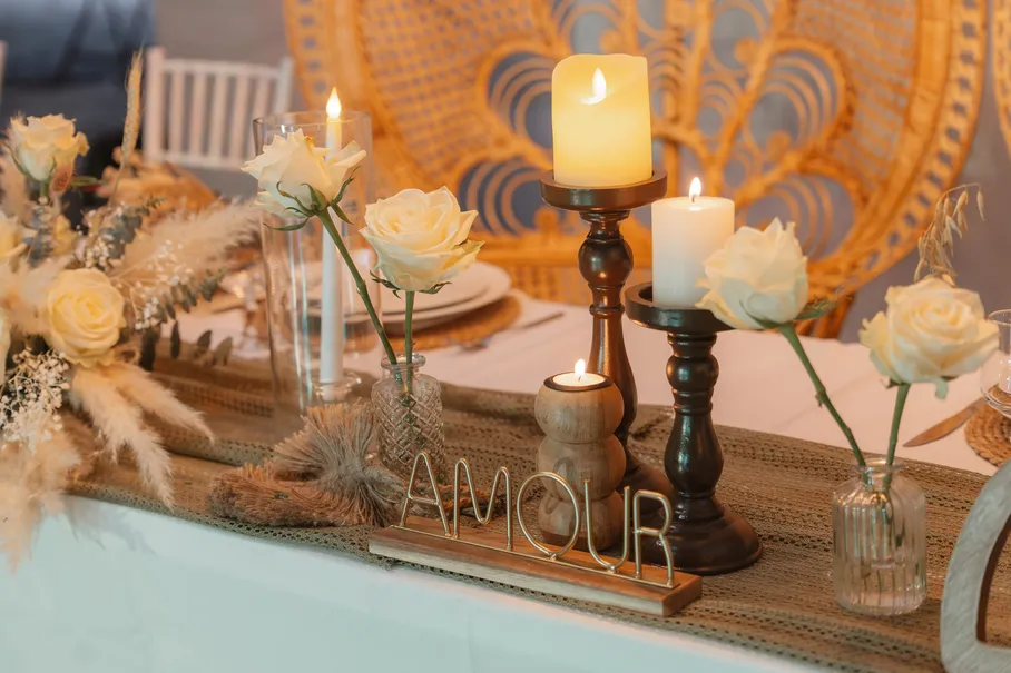 Table décorée de bougies, roses blanches, herbes de la pampa et lettres AMOUR devant un fauteuil en rotin