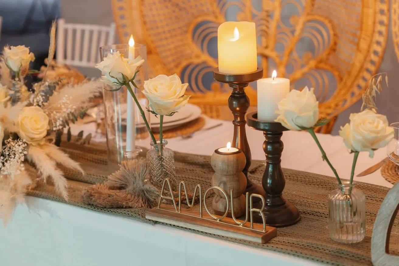 Table décorée de bougies, roses blanches, herbes de la pampa et lettres AMOUR devant un fauteuil en rotin