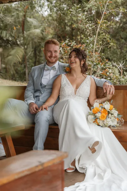 Les mariés assis sur un banc de bois dans la verdure, la mariée riant aux éclats