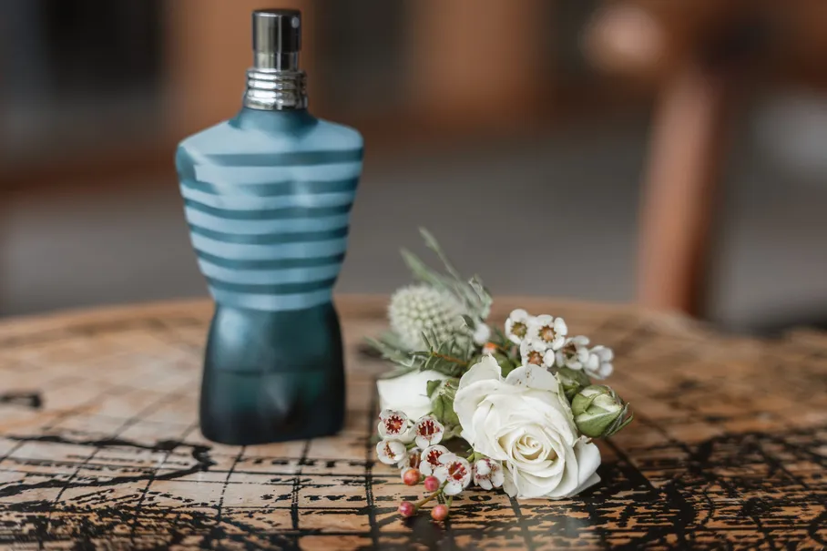 Flacon de parfum bleu rayé à côté d’une boutonnière de roses blanches, sur une table de bois