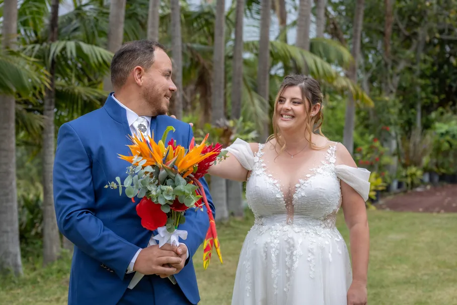 Le marié tient le bouquet coloré et regarde la mariée qui rit, dans un jardin tropical