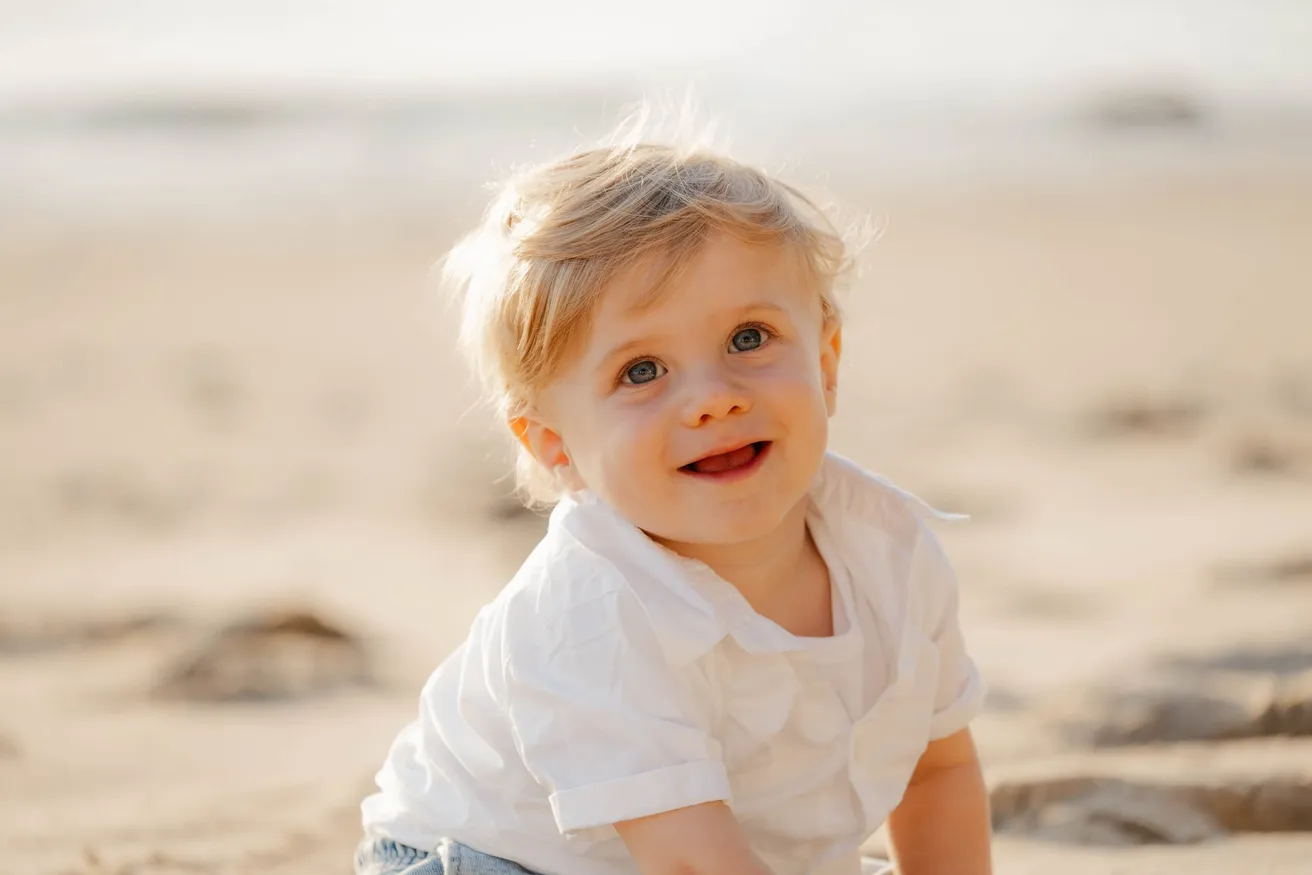 Un petit garçon blond en chemise blanche sourit, assis sur le sable doré
