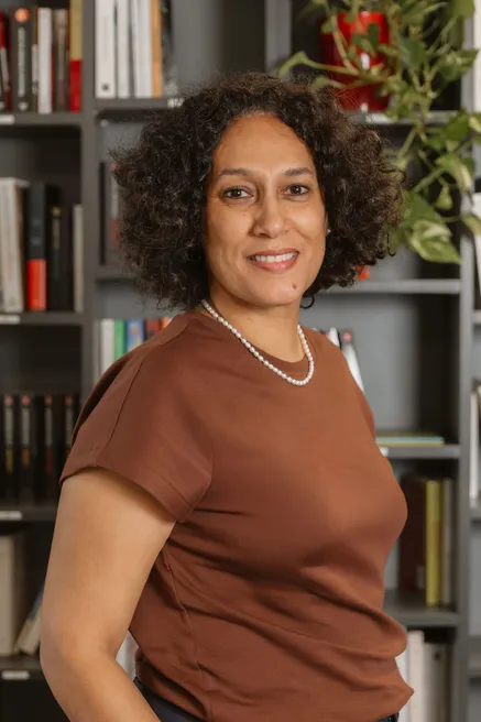 Une femme aux cheveux bouclés, en tee-shirt marron et collier de perles, sourit devant une bibliothèque