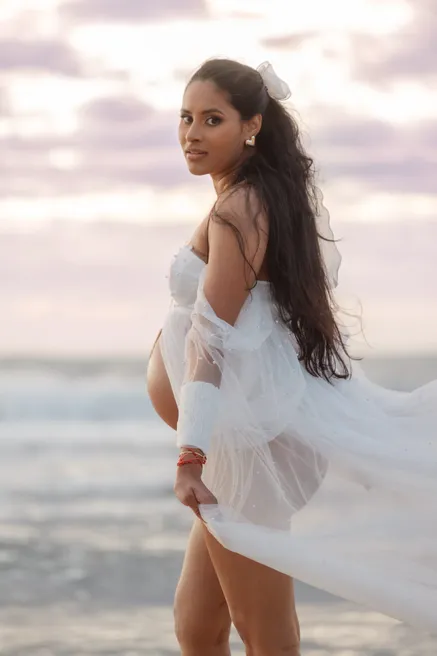 Future maman de dos qui se retourne, voile de tulle blanc porté par le vent, ciel mauve