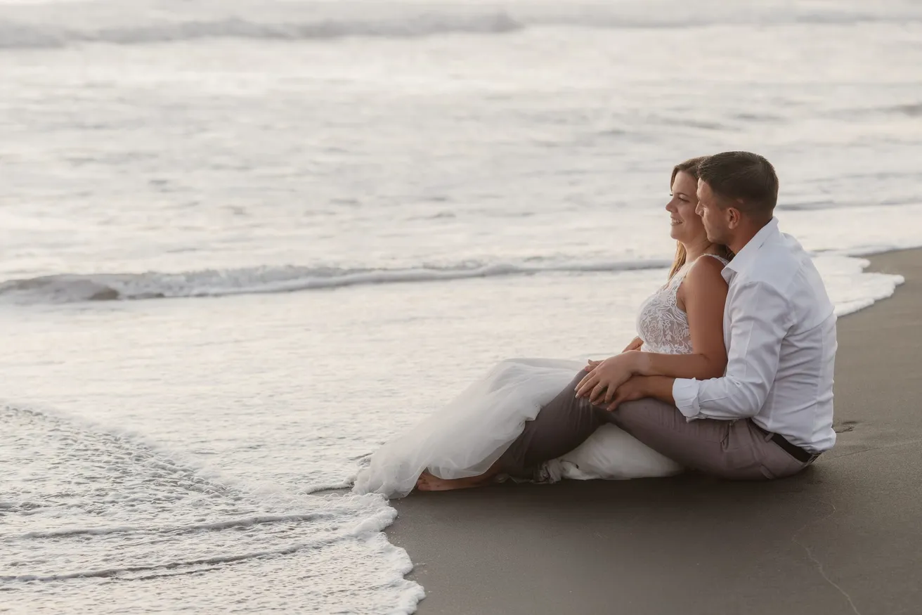 Les mariés assis sur le sable noir au bord des vagues, robe étalée dans l’écume