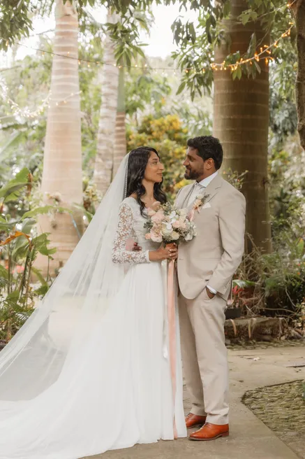 Les mariés côte à côte dans un jardin tropical, la mariée en voile long et dentelle