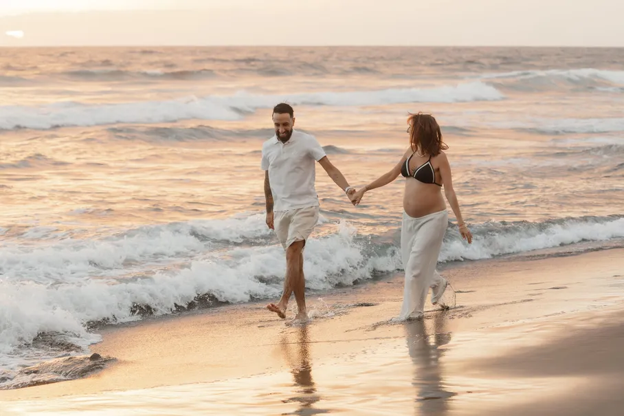 Un couple marche main dans la main au bord des vagues, dans la lumière rosée du soir