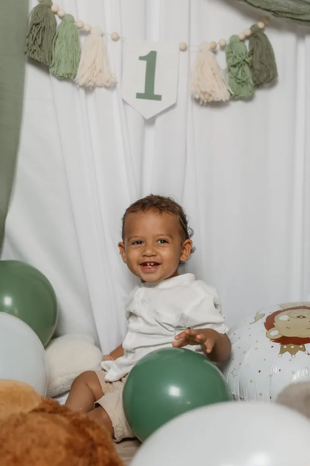 Un petit garçon en chemise blanche sourit, assis au milieu des ballons vert sauge, sous la guirlande « 1 »