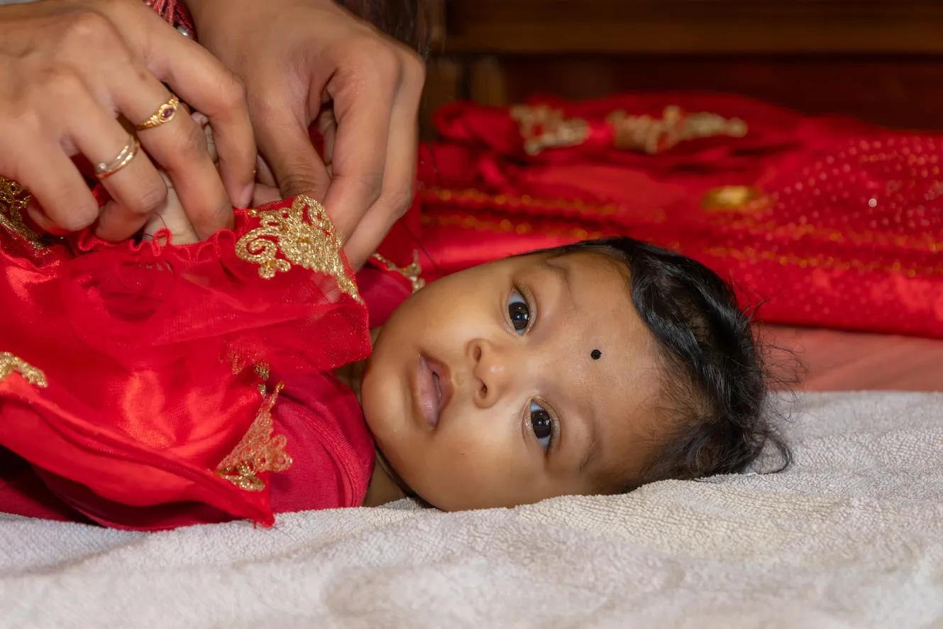 Un bébé en tenue rouge brodée d’or, allongé sur un drap blanc, regarde l’objectif pendant qu’on ajuste son vêtement