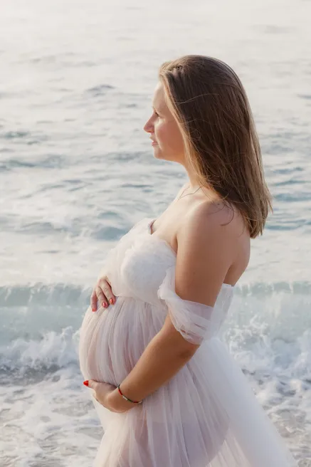 Future maman en robe de tulle, épaules nues, de profil face à la mer, mains sur le ventre