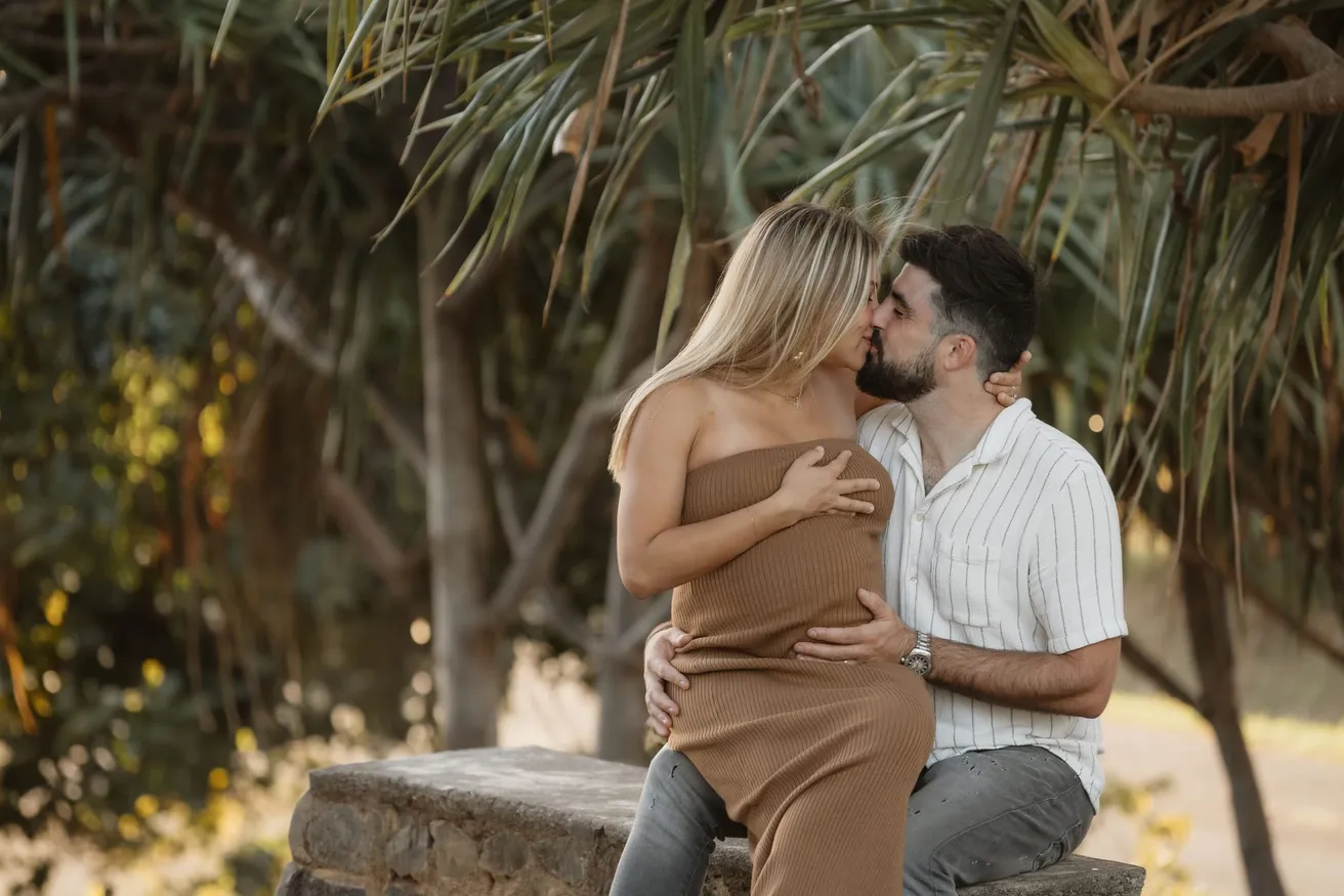Un couple s’embrasse, assis sur un muret à l’ombre des vacoas, la main sur le ventre rond