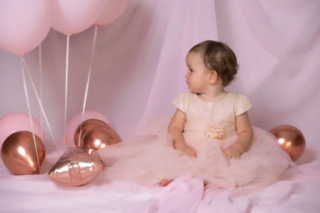 Une petite fille en robe de tulle rose, assise sur le voile, se retourne vers les ballons roses et cuivrés