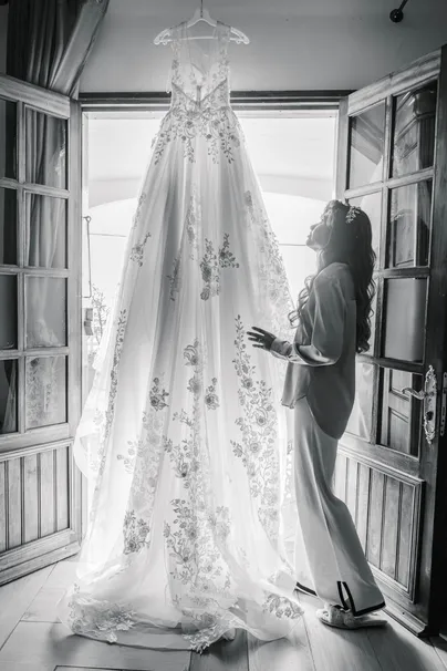En noir et blanc, la robe de mariée suspendue à une porte-fenêtre, la mariée à côté
