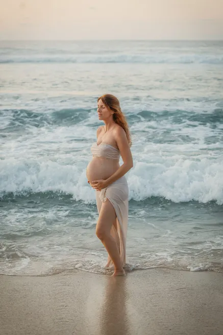 Future maman drapée de blanc debout dans l’écume, face à la houle
