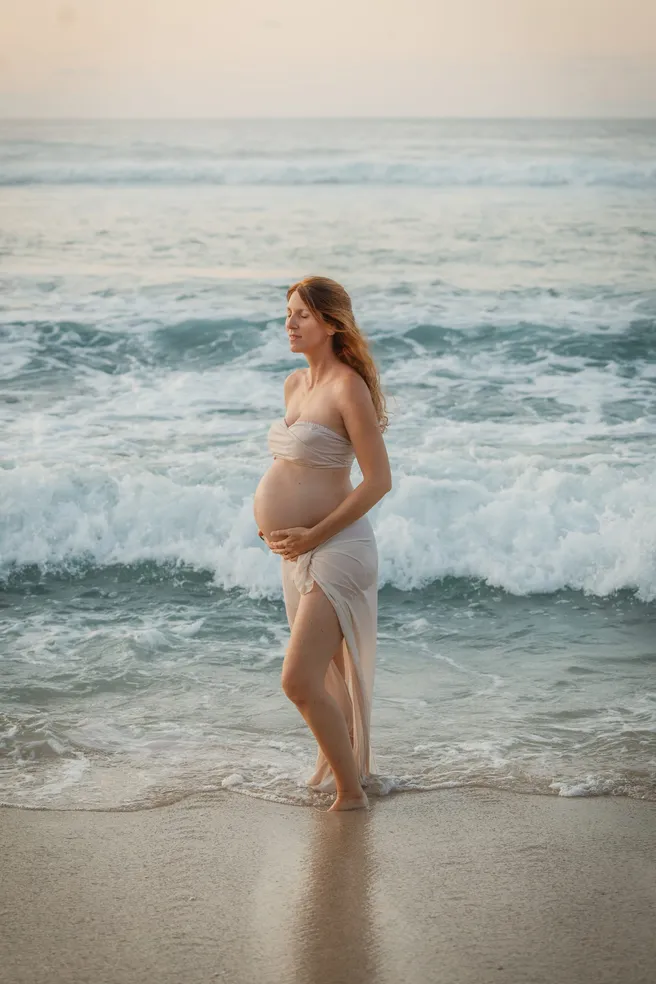 Future maman drapée de blanc debout dans l’écume, face à la houle